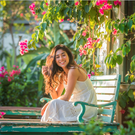 A young woman in a white summer dress sits on a green wooden bench, smiling, surrounded by lush greenery and vibrant pink flowers in a sunlit Elementor-inspired garden.